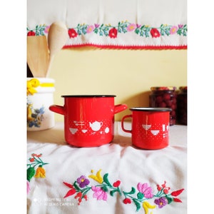 Op de afbeelding: Twee rode emaille potten met witte ontwerpen van keukengerei. De grotere pot heeft een handvat aan elke kant en de kleinere pot heeft een enkel handvat. Beide potten hebben een zwarte rand.