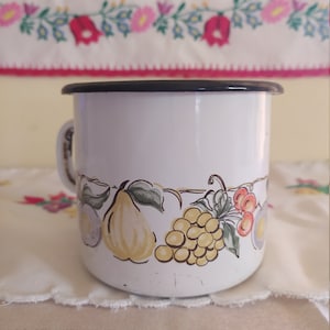 Puede incluir: Taza de esmalte blanco con borde negro y un diseño floral con uvas, peras y cerezas. El diseño está en tonos de amarillo, verde, rojo y morado.