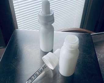 May include: Two white glass bottles with droppers. The bottles are on a silver surface. One bottle has a dropper attached, and the other bottle has the dropper lying next to it. The dropper has markings in milliliters.