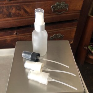 May include: Three frosted glass bottles with different spray tops. The bottles are on a silver surface. The largest bottle has a silver pump top. The smaller bottles have black and white spray tops.