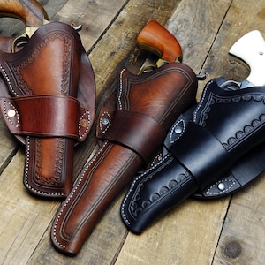 Könnte beinhalten: Drei Leder-Pistolenholster in verschiedenen Braun- und Schwarztönen werden auf einer Holzoberfläche präsentiert. Die Holster weisen dekorative Nähte und geprägte Muster auf. Jedes Holster hat einen Riemen zur Sicherung der Waffe.