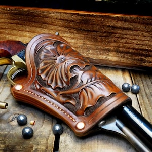 May include: A brown leather holster featuring an intricate floral carving pattern, secured with copper-colored rivets. The holster holds a firearm with a dark barrel and brass accents, accompanied by several small round lead balls nearby.