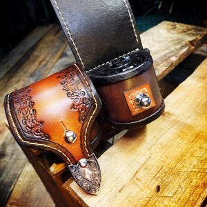 May include: A brown leather camera strap with a decorative design. The strap has a metal buckle and a brown leather pouch with a metal button.