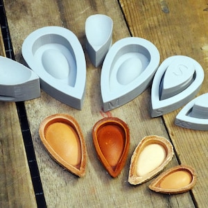 May include: A collection of gray, teardrop-shaped molds and several open almond shells in varying shades of brown and tan. The molds are arranged on a wooden surface, suggesting a craft or food preparation theme.