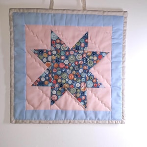 May include: A square quilted wall hanging with a star design. The star is made of fabric patterned with colourful buttons on a blue background. The star is set against a pink background, framed by a light blue border and a beige edge.