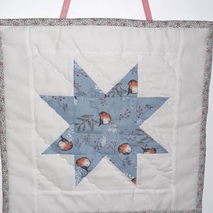 May include: A square quilted wall hanging with a central blue star design, adorned with robin illustrations and floral details. The star is set against a white backdrop, framed by a floral border and a pink ribbon for hanging.