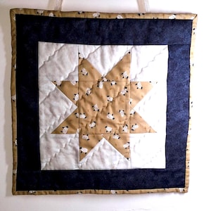 Op de afbeelding: Een gewatteerde wandhanger met een sterontwerp. De ster is gemaakt van bruin textiel met een schapenpatroon, tegen een witte en marineblauwe achtergrond. De randen zijn afgewerkt met een patroonrand en een crèmekleurig lint om op te hangen.