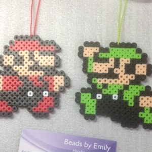 May include: Two pixel art ornaments of Mario and Luigi characters made with colorful beads. The ornaments are hanging from red and green strings.