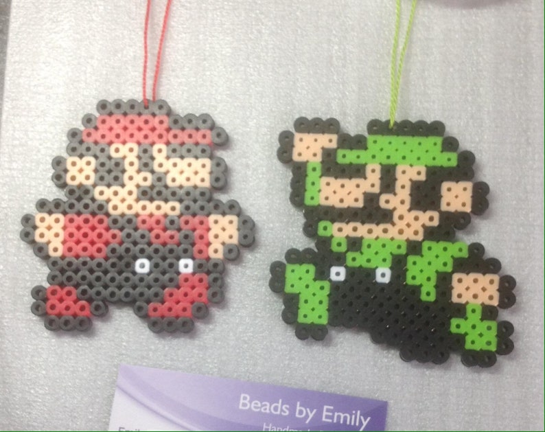 May include: Two pixel art ornaments of Mario and Luigi characters made with colorful beads. The ornaments are hanging from red and green strings.