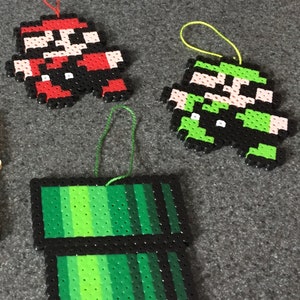 May include: Three handmade Christmas ornaments featuring characters from the Super Mario Bros. video game. The ornaments are made of colorful plastic beads and feature Mario, Luigi, and a green pipe.