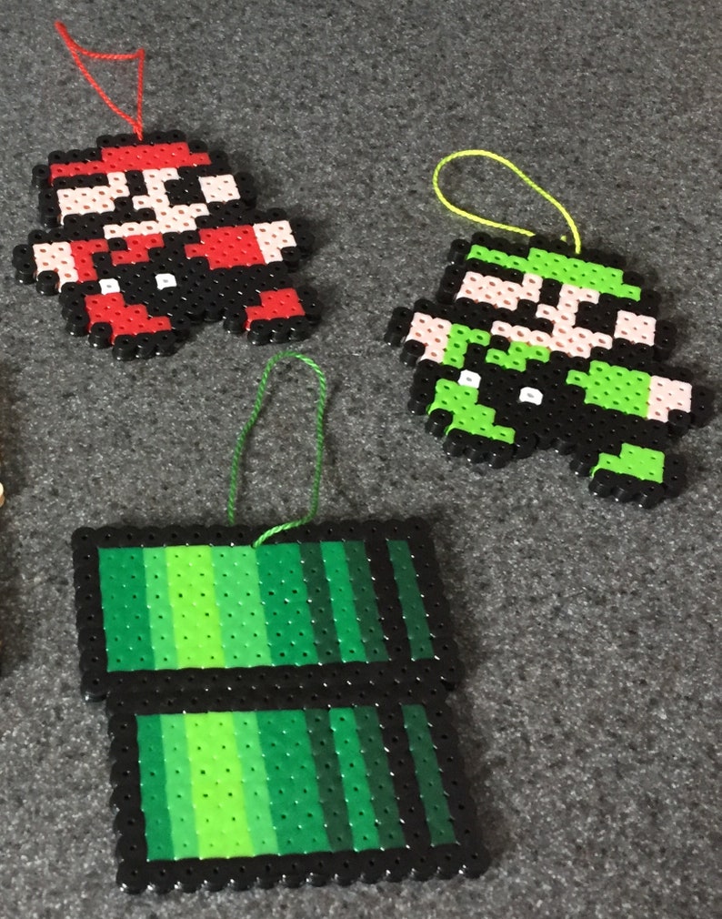 May include: Three handmade Christmas ornaments featuring characters from the Super Mario Bros. video game. The ornaments are made of colorful plastic beads and feature Mario, Luigi, and a green pipe.