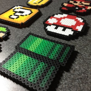 May include: A set of pixel art characters and items from the Super Mario Bros. video game. The characters and items are made of colorful plastic beads and are arranged on a gray surface. The characters include Mario, a question block, a coin, a mushroom, and a green pipe.