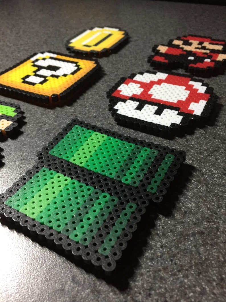 May include: A set of pixel art characters and items from the Super Mario Bros. video game. The characters and items are made of colorful plastic beads and are arranged on a gray surface. The characters include Mario, a question block, a coin, a mushroom, and a green pipe.