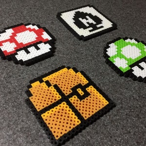 May include: Four pixel art designs made with colorful beads. The designs include a red and white mushroom, a green and white mushroom, a yellow and orange brick block, and a white and black block with a question mark.