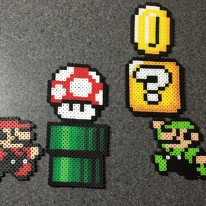 May include: A set of five pixel art characters from the Super Mario Bros. video game series. The characters include Mario, Luigi, a mushroom, a question block, and a coin. The characters are made of colorful beads.