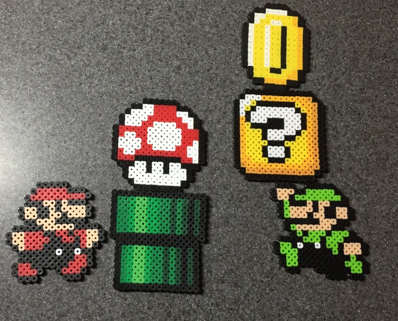 May include: A set of five pixel art characters from the Super Mario Bros. video game series. The characters include Mario, Luigi, a mushroom, a question block, and a coin. The characters are made of colorful beads.