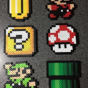 May include: A set of six pixel art characters from the Super Mario Bros. video game series. The characters are made of colorful plastic beads and include Mario, Luigi, a mushroom, a coin, a question block, and a green pipe.