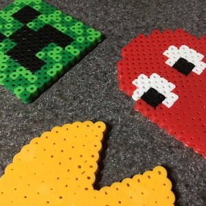 May include: Three pixel art characters made with colorful beads. The characters are Pac-Man, a ghost, and a green creeper from the video game Minecraft.