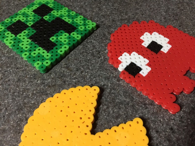 May include: Three pixel art characters made with colorful beads. The characters are Pac-Man, a ghost, and a green creeper from the video game Minecraft.