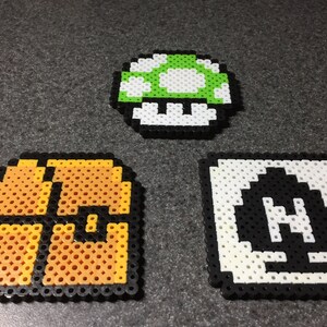 May include: Three pixel art crafts made with beads. A green and white mushroom, a gold treasure chest, and a black and white pixel art of the letter H.