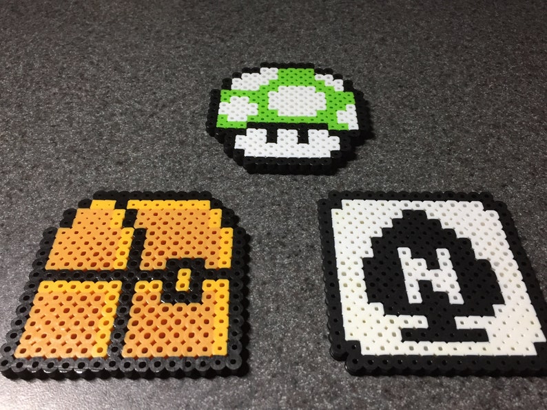 May include: Three pixel art crafts made with beads. A green and white mushroom, a gold treasure chest, and a black and white pixel art of the letter H.