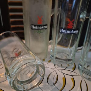 Puede incluir: Varias jarras de cerveza de vidrio transparente, algunas escarchadas, con asas, se exhiben en una bandeja decorativa. Las jarras presentan el logotipo de Heineken con un molino de viento rojo y el nombre de la marca en negro. Una jarra est&aacute; tumbada.