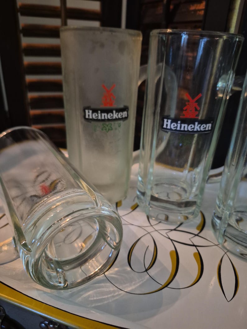 Puede incluir: Varias jarras de cerveza de vidrio transparente, algunas escarchadas, con asas, se exhiben en una bandeja decorativa. Las jarras presentan el logotipo de Heineken con un molino de viento rojo y el nombre de la marca en negro. Una jarra est&aacute; tumbada.
