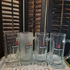 Puede incluir: Cuatro jarras de cerveza de vidrio transparente con asas y una jarra esmerilada, todas con el logotipo "Heineken". Las jarras est&aacute;n sobre una bandeja decorativa con un dise&ntilde;o blanco y dorado, sobre un fondo de persianas de madera oscura.