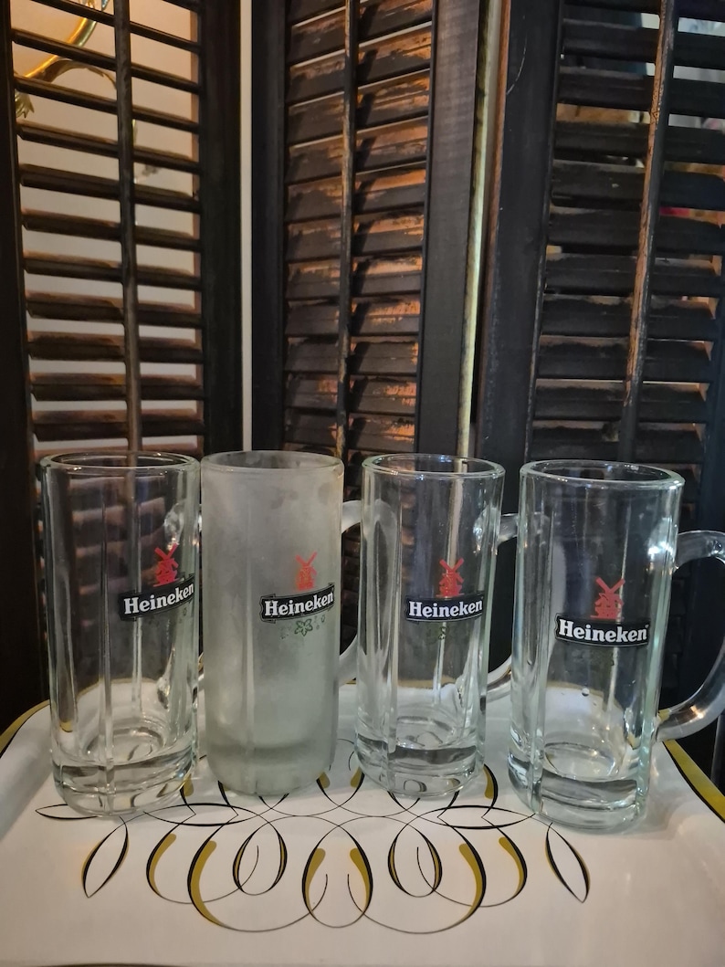 Puede incluir: Cuatro jarras de cerveza de vidrio transparente con asas y una jarra esmerilada, todas con el logotipo "Heineken". Las jarras est&aacute;n sobre una bandeja decorativa con un dise&ntilde;o blanco y dorado, sobre un fondo de persianas de madera oscura.