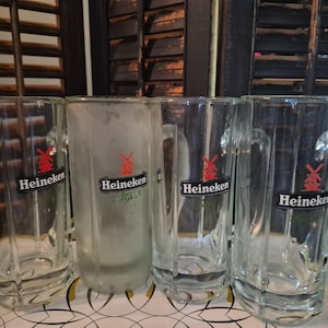 Puede incluir: Cuatro jarras de cerveza de vidrio transparente con asas, cada una con el logotipo de Heineken con un molino de viento rojo. Una jarra est&aacute; esmerilada. Las jarras est&aacute;n dispuestas sobre una bandeja blanca, con un fondo de contraventanas de madera oscura.
