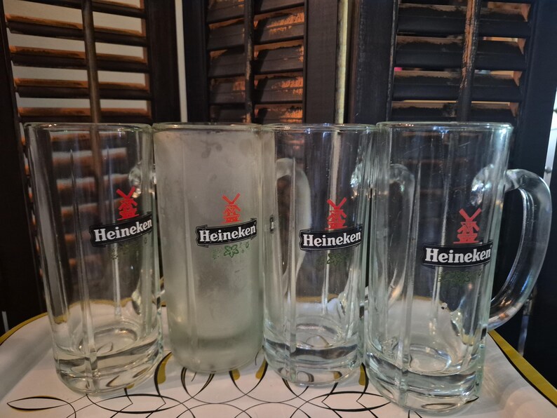 Puede incluir: Cuatro jarras de cerveza de vidrio transparente con asas, cada una con el logotipo de Heineken con un molino de viento rojo. Una jarra est&aacute; esmerilada. Las jarras est&aacute;n dispuestas sobre una bandeja blanca, con un fondo de contraventanas de madera oscura.
