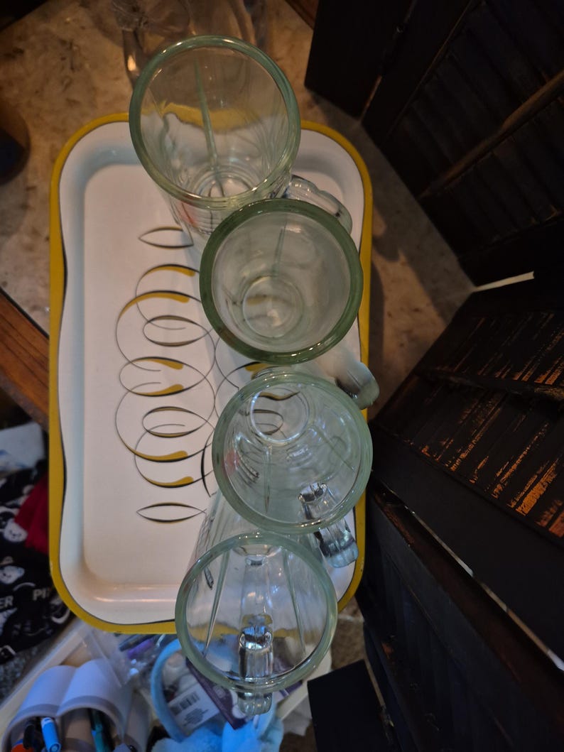 Puede incluir: Cuatro tazas de vidrio transparente con asas est&aacute;n dispuestas sobre una bandeja blanca con un borde amarillo y un dise&ntilde;o retro. Las tazas tienen un borde verde. La bandeja est&aacute; sobre una superficie con otros elementos en el fondo.