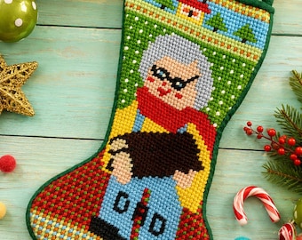 May include: A needlepoint Christmas stocking featuring a colourful design of a person with grey hair, glasses, and a red scarf, holding a dark object. The background shows a small house and green trees, with a green fabric border and hanging loop.