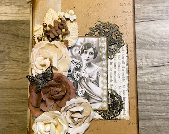Puede incluir: Un álbum de recortes de estilo vintage con una cubierta de cuero marrón y adornos decorativos. La cubierta presenta un diseño floral con flores de papel marrón y blanco, una mariposa y una fotografía vintage de una mujer. El álbum tiene un cierre de metal dorado y acentos de esquina.