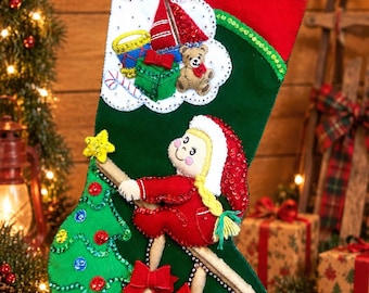 May include: A green felt Christmas stocking featuring a red cuff and a decorative applique of a blonde character in a red outfit climbing a ladder toward a star-topped tree. The design includes a white cloud shape with toys and sequins.