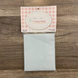 May include: A package of Vintage Cloth cross-stitch fabric. The fabric is light blue and has a grid pattern. The packaging has a pink and white gingham pattern and the text "Vintage Cloth" and "18" x 21" Breeze".