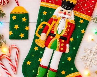 May include: A green and red felt Christmas stocking featuring a central nutcracker figure holding a gold trumpet, decorated with sequins and yellow stars, accompanied by a matching rectangular felt gift tag with yellow stars on a green background.