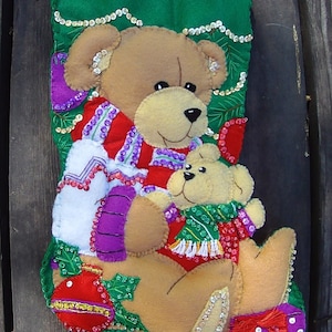 May include: A Christmas stocking made of green felt, decorated with a large brown teddy bear holding a smaller one. The bears wear red and white outfits and green scarves. The stocking is embellished with sequins and a red ornament.