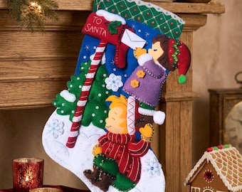 May include: A felt Christmas stocking featuring a blue background with a green cuff, a red mailbox labelled TO SANTA, and two figures. One figure in a purple coat places a letter in the mailbox, while another in a red jumper stands below.