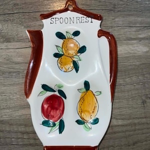 May include: A white ceramic spoon rest shaped like a coffee pot with a brown rim and handle. The text "SPOON REST" is printed at the top. The surface features hand-painted illustrations of two orange citrus fruits, a red apple, and a yellow pear.