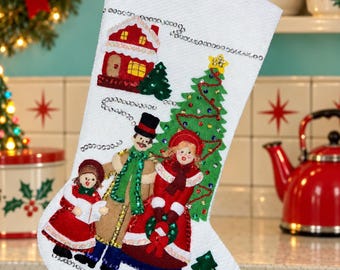 May include: A white felt Christmas stocking featuring an appliqued scene of a family caroling near a decorated green Christmas tree and a small house. The design includes a small dog, sequin accents, and embroidered details throughout the fabric.