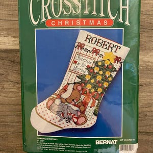May include: A Christmas cross-stitch kit with a stocking design. The stocking features a Christmas tree, teddy bears, and the name "Robert". The kit includes fabric, floss, needles, and instructions. The package is green and reads "Cross Stitch Christmas". The stocking measures 23 cm x 41 cm.
