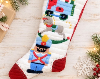 May include: A white quilted Christmas stocking featuring embroidered designs of a blue and red toy train, a grey rocking horse, and a blue nutcracker soldier. The stocking has a red toe, a red heel, and a red hanging loop at the top edge.