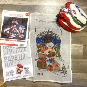 May include: A hand-painted needlepoint Christmas stocking kit featuring a snowman design, with a bundle of colorful yarn. The kit includes a canvas stocking, instructions, and a chart for personalization. The stocking is approximately 18 inches long.