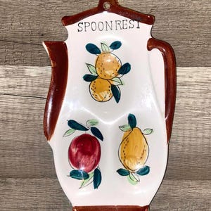 May include: White ceramic spoon rest with brown trim and handle. The spoon rest is decorated with hand-painted fruit illustrations, including oranges, a red apple, and a yellow pear. The words "SPOON REST" are printed at the top.