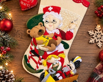 May include: A cream-coloured felt Christmas stocking featuring an appliqued nurse figure holding a teddy bear, a small doll, and a nutcracker in a toy wagon. A red heart-shaped ornament attached to the top reads, Nursing is a work of Heart!