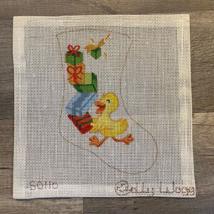 May include: Needlepoint canvas featuring a yellow duck carrying gifts. The gifts are green, red, and blue. The canvas has the words "Soho" and "Baby Daze" in the corners. The background is a white grid.