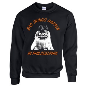 Könnte beinhalten: Schwarzer Pullover mit einem weißen Grafikdruck eines pelzigen Wesens, das einen Hut trägt. Der Text "Bad Things Happen in Philadelphia" ist in Orange gedruckt.