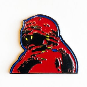 May include: Red enamel pin with a black and yellow creature with sharp claws and teeth. The creature is wearing a red hooded cloak with a blue outline.