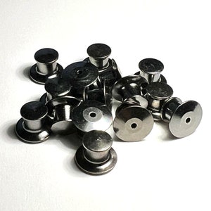 May include: A pile of silver metal pin backs for crafts and jewelry making. The pin backs are round and have a small hole in the center.